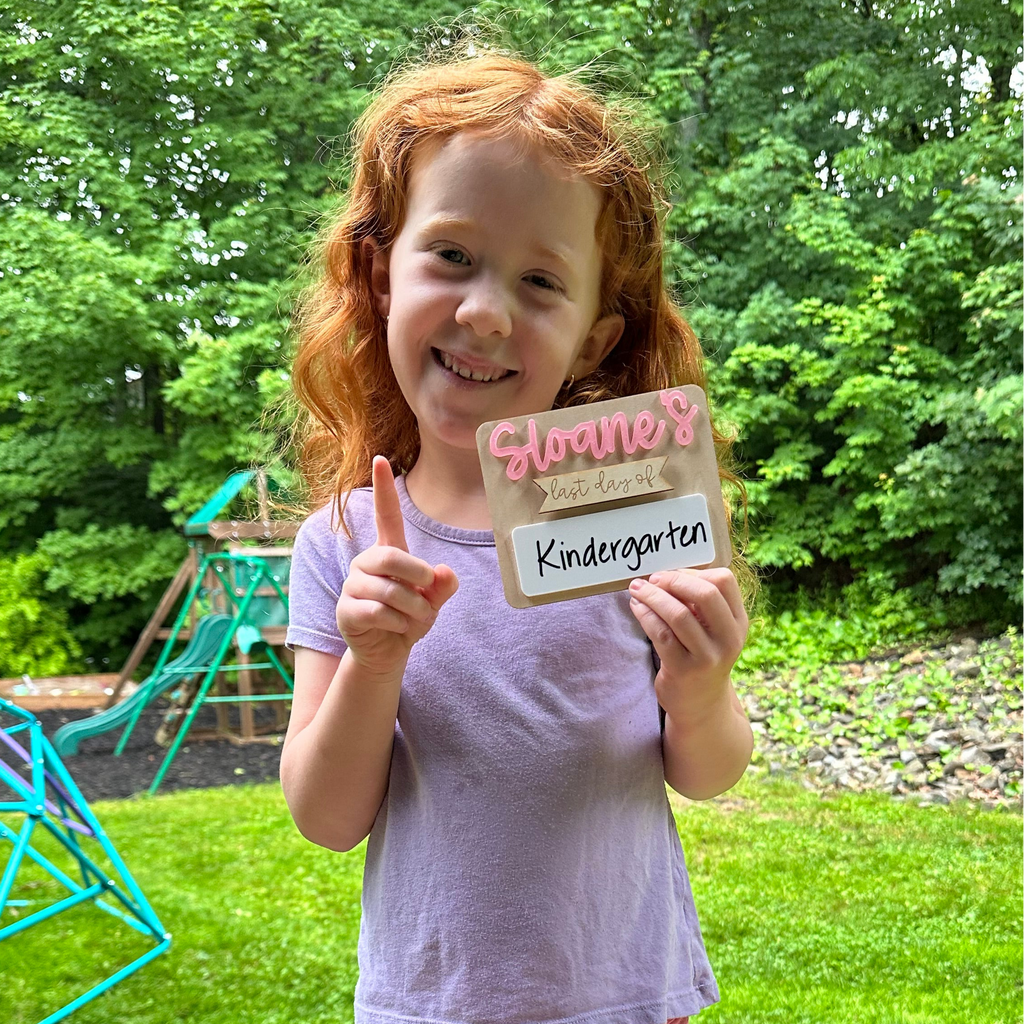 Young girl holding a sign with 'Sloane's last day of Kindergarten' outdoors.